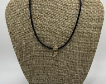 May include: A black cord necklace with a gold-colored clasp and a small, curved pendant. The pendant is embellished with small, sparkling details. The necklace is displayed on a neutral-toned mannequin.