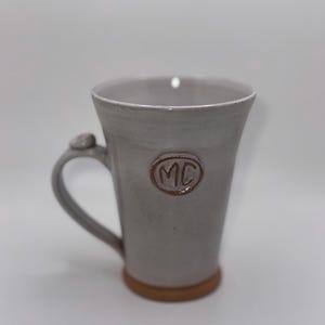 May include: A gray ceramic mug with a brown base and handle. The mug has a flared shape and features the initials "MC" in a circular design. The handle has a small, decorative element. The mug is suitable for coffee, tea, or other beverages.