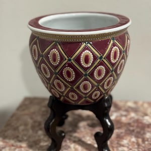 May include: A decorative burgundy ceramic planter with a geometric pattern of gold and white ovals and diamonds. The planter sits on a dark wooden stand with three curved legs. The rim of the planter is trimmed with gold.
