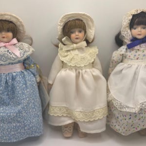 Può includere: Tre bambole di porcellana in abiti vintage. Le bambole hanno volti dipinti e indossano cappellini. Gli abiti sono blu, bianchi e a fantasia floreale, con dettagli in pizzo e nastri. Le bambole sono in piedi fianco a fianco.