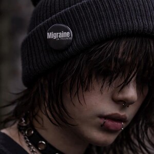 May include: A dark grey beanie with a button that says "Migraine IN PROGRESS". The person is wearing a black choker necklace and has multiple facial piercings. The person has dark hair.