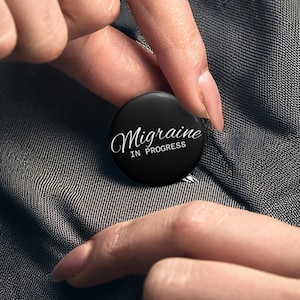 Op de afbeelding: Een zwarte knop met de woorden "Migraine IN PROGRESS" in witte letters. De knop wordt tussen vingers gehouden tegen een grijze stoffen achtergrond. De knop is rond en glanzend.