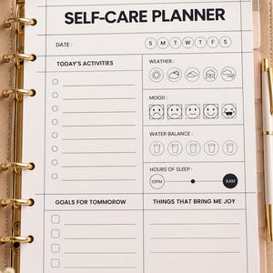 May include: A beige ring-bound self-care planner with a white page featuring sections for date, activities, weather, mood, water balance, sleep hours, goals for tomorrow, and things that bring joy, accompanied by a gold pen.