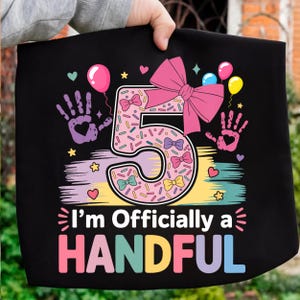 Puede incluir: Camiseta negra con un diseño colorido para un quinto cumpleaños. El número 5 está decorado con chispas y lazos, con el texto "I'm Officially a HANDFUL" en rosa y azul.