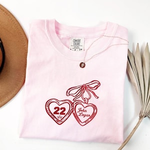 May include: A light pink Comfort Colors t-shirt with a red embroidered design of two heart-shaped lockets, one with the number "22" and a crown, the other with the name "John Logan". A delicate necklace with a round pendant is draped on the shirt.