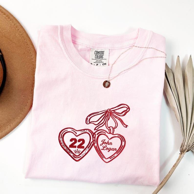 May include: A light pink Comfort Colors t-shirt with a red embroidered design of two heart-shaped lockets, one with the number "22" and a crown, the other with the name "John Logan". A delicate necklace with a round pendant is draped on the shirt.
