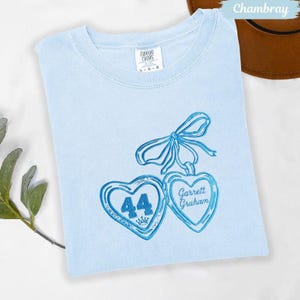 May include: Light blue t-shirt with embroidered blue heart pendants. One heart has the number 44 and a crown, the other says "Garrett Graham". The shirt is a Comfort Colors brand.