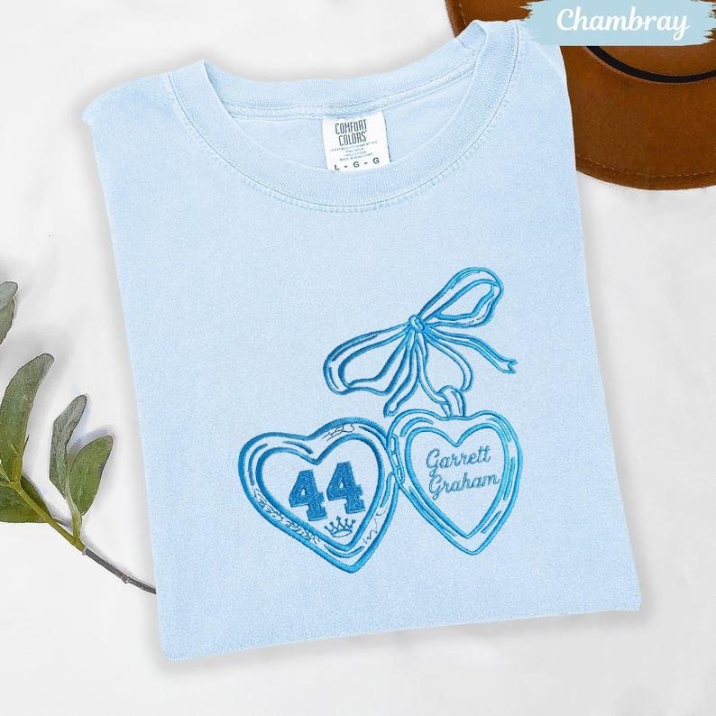 May include: Light blue t-shirt with embroidered blue heart pendants. One heart has the number 44 and a crown, the other says "Garrett Graham". The shirt is a Comfort Colors brand.