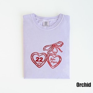May include: Orchid-colored t-shirt with a red heart design. The design features two heart-shaped pendants with the number "22" and the name "John Logan". A red bow connects the pendants. The shirt is a size XL-TG-EG.