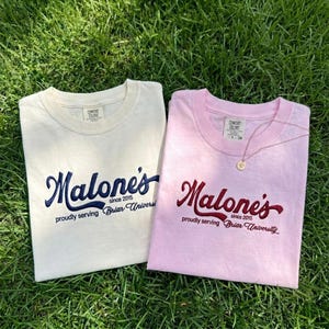 May include: Two folded t-shirts on grass. One is cream-colored with navy blue script that reads "Malone's since 2015 proudly serving Briar University." The other is pink with burgundy script. A gold necklace rests on the pink shirt.