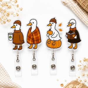 May include: Four acrylic badge reels featuring cartoon ducks in autumn themes. One duck wears a brown sweater and holds a coffee cup, one is wrapped in a plaid blanket, one sits on a pumpkin, and one wears glasses while reading a book.