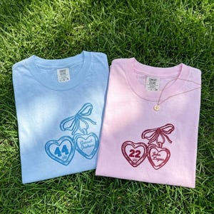 May include: Two Comfort Colors t-shirts, one light blue and one pink, are displayed on green grass. Each shirt features a heart design with a bow, number, and name. The light blue shirt has the number 44 and the name "Garrett Graham". The pink shirt has the number 22 and the name "John Logan".