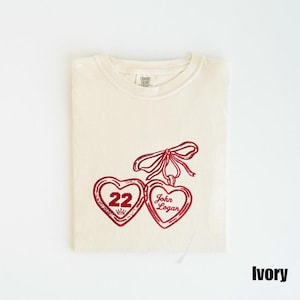 May include: Ivory-colored t-shirt with a red heart design. One heart has the number 22 and a crown, the other says "John Logan". A red bow connects the hearts. The shirt is folded neatly.