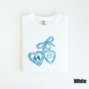 May include: White t-shirt with a blue heart design. The design includes two heart-shaped pendants, one with the number 44 and a crown, and the other with the name "Garrett Graham". A blue bow is at the top.