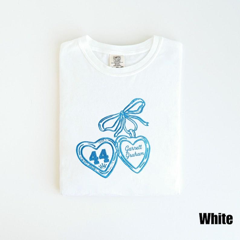 May include: White t-shirt with a blue heart design. The design includes two heart-shaped pendants, one with the number 44 and a crown, and the other with the name "Garrett Graham". A blue bow is at the top.