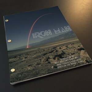 Può includere: Una copertina di sceneggiatura blu scuro con il titolo "IRON MAN" in lettere bianche bordate. La copertina presenta un arco rosso e bianco nel cielo sopra un paesaggio desertico. Il testo "SCREENPLAY BY MATT HOLLOWAY & ART MARCUM MARK FERGUS & HAWK OSTBY BASED ON THE MARVEL COMIC" è stampato in bianco nella parte inferiore della copertina.