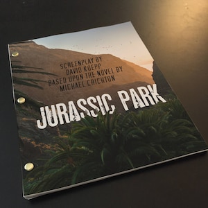 May include: A close-up of the front cover of a Jurassic Park screenplay. The cover features a scenic image of a lush jungle with a mountain range in the background. The title "Jurassic Park" is printed in white with a distressed effect. The text "Screenplay by David Koepp Based upon the novel by Michael Crichton" is printed in white below the title.