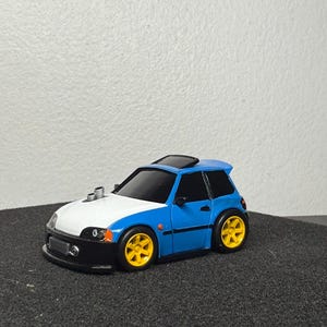 Op de afbeelding: Een kleine, gestileerde blauw-witte speelgoedauto met gele wielen. De auto heeft een zwarte voorbumper en een zonnedak. Het ontwerp omvat uitlaatpijpen en een sportieve uitstraling.