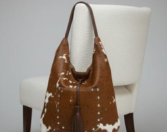 May include: A brown and white cowhide hobo bag with a leather handle and a tassel. The bag has silver-tone stud accents and is draped over a white upholstered chair.