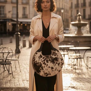 Artisanal round cowhide crossbody bag with premium tri-color brown, black, and white hair-on-hide. This circular western purse features hand-sewn black leather whipstitch edges and a structured top handle. A unique boho-chic statement handbag for rodeo fashion, country weddings, or a luxury bohemian wardrobe.