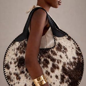 Handcrafted round cowhide tote bag with authentic tri-color brown, black, and white hair-on-hide texture. The circular hobo silhouette features hand-stitched black leather trim and a top handle, showcased in a sophisticated home office setting with a vintage aesthetic.