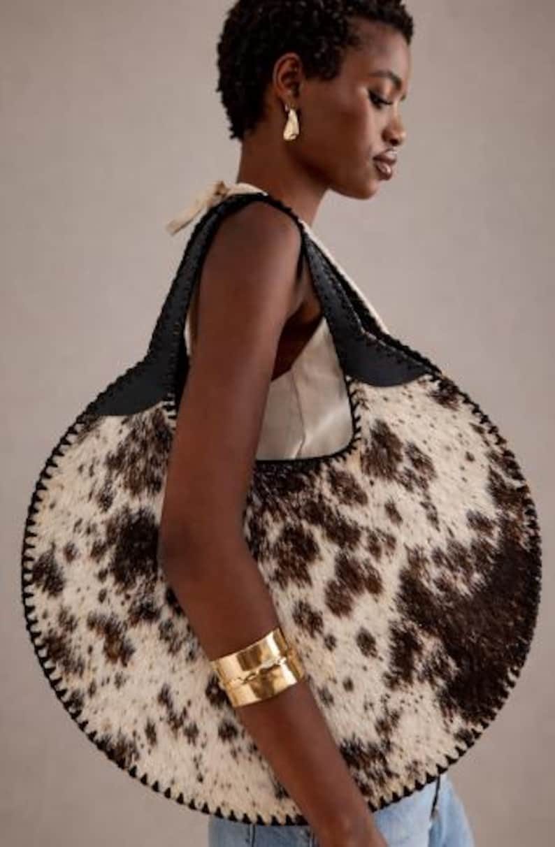 Handcrafted round cowhide tote bag with authentic tri-color brown, black, and white hair-on-hide texture. The circular hobo silhouette features hand-stitched black leather trim and a top handle, showcased in a sophisticated home office setting with a vintage aesthetic.