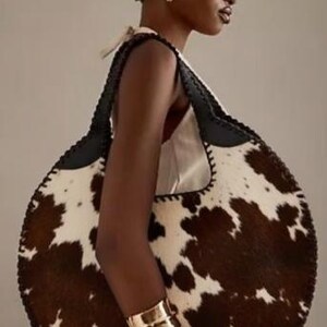 May include: A large, round handbag with a cowhide pattern in brown and white. The bag has a black leather strap and trim. A gold cuff bracelet is visible on the arm.