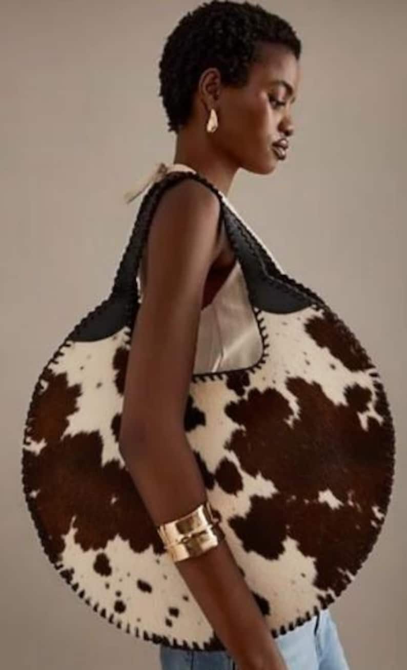 May include: A large, round handbag with a cowhide pattern in brown and white. The bag has a black leather strap and trim. A gold cuff bracelet is visible on the arm.