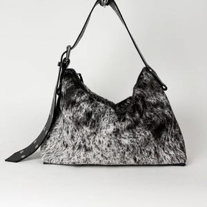May include: A hobo-style shoulder bag featuring a black and white speckled animal print texture. The bag includes a black leather strap with silver-toned metal hardware, including a buckle and rivets, attached to the top of the bag structure.