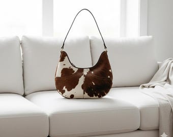 May include: A brown and white cowhide shoulder bag with a black strap. The bag is crescent-shaped and sits on a white couch. The bag's pattern is a mix of brown and white patches.