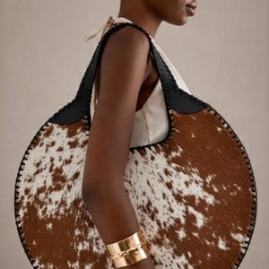 May include: A large, round handbag with a cowhide pattern in brown and white. The bag has black leather straps and trim. A gold cuff bracelet is visible on the arm.
