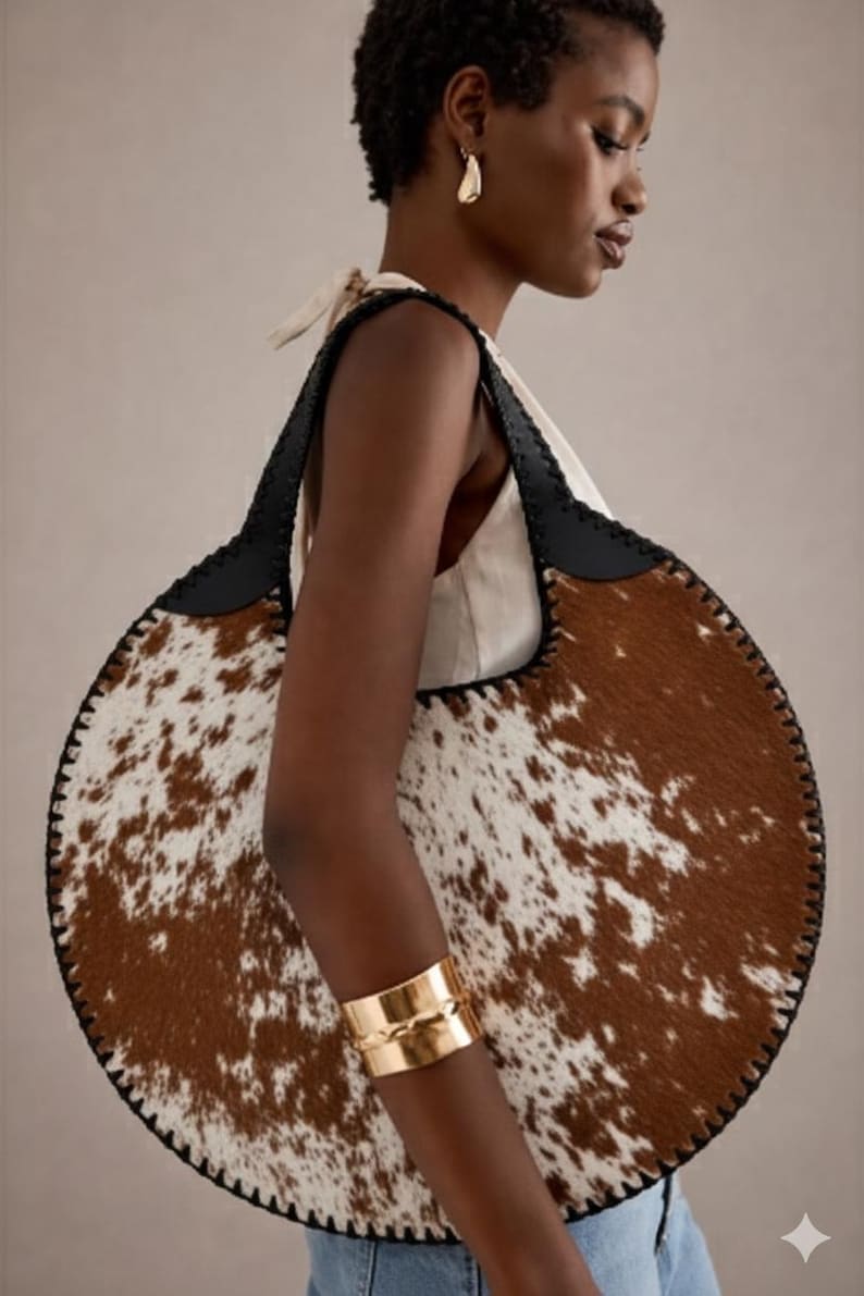 May include: A large, round handbag with a cowhide pattern in brown and white. The bag has black leather straps and trim. A gold cuff bracelet is visible on the arm.