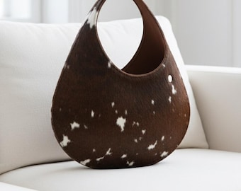 May include: A brown and white cowhide handbag with a single handle. The bag has a teardrop shape and a brown interior. A silver button is visible on the side of the bag. The bag is sitting on a white sofa.