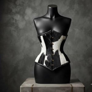 May include: A corset featuring a black and white cow print pattern with a textured finish. The garment has a front closure with a vertical row of small metallic buttons and is displayed on a black mannequin torso, showcasing a fitted silhouette.