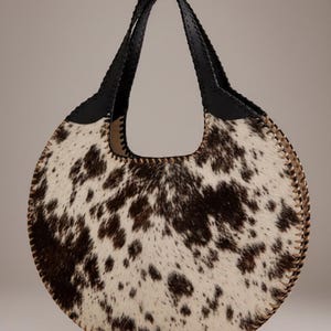 May include: A round handbag with a cowhide pattern in shades of brown and white. The bag has a black leather handle and trim, and a stitched edge. The bag is designed with a circular shape and a large opening.