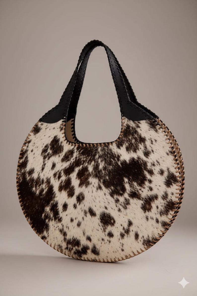 May include: A round handbag with a cowhide pattern in shades of brown and white. The bag has a black leather handle and trim, and a stitched edge. The bag is designed with a circular shape and a large opening.