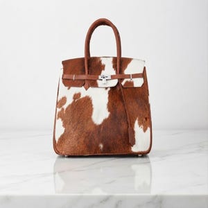May include: A structured handbag featuring a brown and white cowhide pattern with a textured fur finish. The bag includes brown leather handles, a horizontal leather strap across the front, and a silver-toned metal lock closure mechanism.