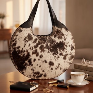 Luxurious round cowhide hobo bag featuring a natural tri-color brown, white, and black speckled pattern. This circular leather handbag includes a structured black leather handle with whipstitch detailing, displayed on a mahogany wooden table alongside elegant lifestyle accessories like a teacup and gold pens.