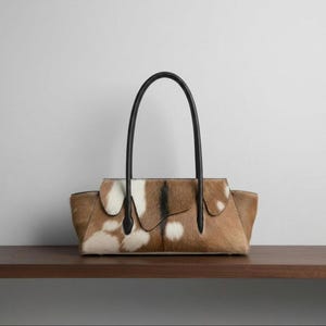 Könnte beinhalten: Eine horizontale Handtasche mit einem braun-weißen Kuhfellmuster und einer strukturierten Felloberfläche. Die Tasche hat eine strukturierte Silhouette mit einer geschwungenen oberen Klappe und zwei dünnen, schwarzen Ledergriffen an den Seiten.
