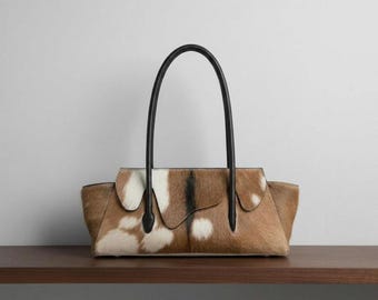 May include: A horizontal handbag featuring a brown and white cowhide pattern with a textured fur finish. The bag has a structured silhouette with a curved top flap and two thin, black leather handles attached to the front and back panels.