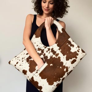 May include: A large, square-shaped handbag with a cowhide pattern in brown and white. The bag has a circular handle and a pocket on the side. The bag is held by a person wearing a black dress.