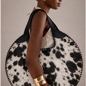 May include: A large, round handbag with a cowhide pattern in black and white. The bag has black leather handles and trim, and is carried over the shoulder. The model is wearing a gold cuff bracelet.