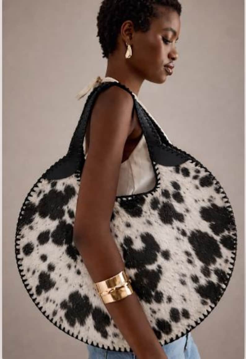 May include: A large, round handbag with a cowhide pattern in black and white. The bag has black leather handles and trim, and is carried over the shoulder. The model is wearing a gold cuff bracelet.