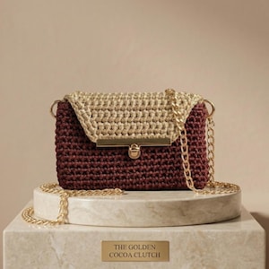 Op de afbeelding: Een handgemaakte, bordeauxrode en beige gehaakte clutch met een gouden ketting. De tas heeft een gouden sluiting en staat op een marmeren sokkel. De tekst op de sokkel luidt "THE GOLDEN COCOA CLUTCH".