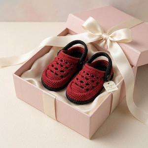 May include: A pair of dark red crocheted baby shoes with black straps and soles, featuring a perforated design. The shoes are presented in a light pink gift box with a satin ribbon and a small white tag that reads Handmade for Little Feet.