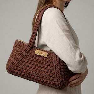 May include: A brown crochet handbag with a rectangular shape and a gold zipper. The bag has two handles and a rectangular label that reads "VERSACE JEANS COUTURE". The bag is held by a person.