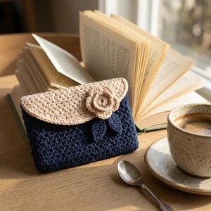 Puede incluir: Un estuche rectangular de ganchillo con un cuerpo principal de color azul marino oscuro y una solapa beige en contraste. La solapa está decorada con una flor de ganchillo tridimensional y dos pequeñas hojas azul marino en el frente.