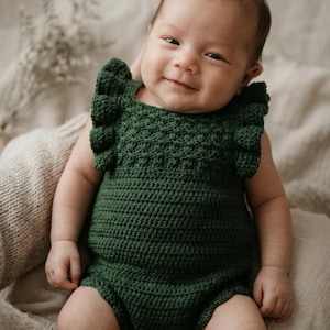 May include: A dark forest green crochet romper featuring a textured bodice with a bobble stitch pattern, ruffled shoulder straps, and three small white buttons at the bottom closure. The garment is crafted with a dense yarn construction.