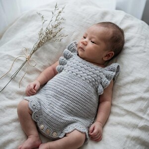 May include: A light gray crocheted romper featuring a textured bodice with ruffled sleeves and a scalloped hemline. The garment includes three small, round, light-colored buttons at the bottom center, providing a functional closure for the leg openings.