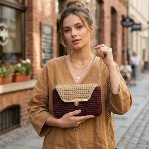 May include: A burgundy and tan crochet handbag with a gold-colored chain strap and clasp. The bag has a rectangular shape with a flap closure. The background shows a brick building and a street.
